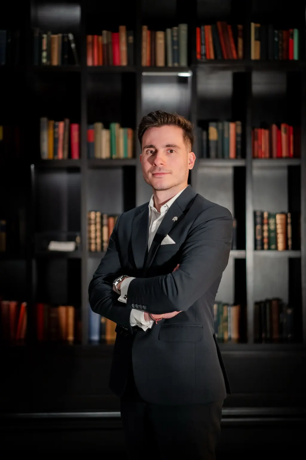 Michalis Kiotis in a dark suit, arms folded, in front of a wall of bookshelves